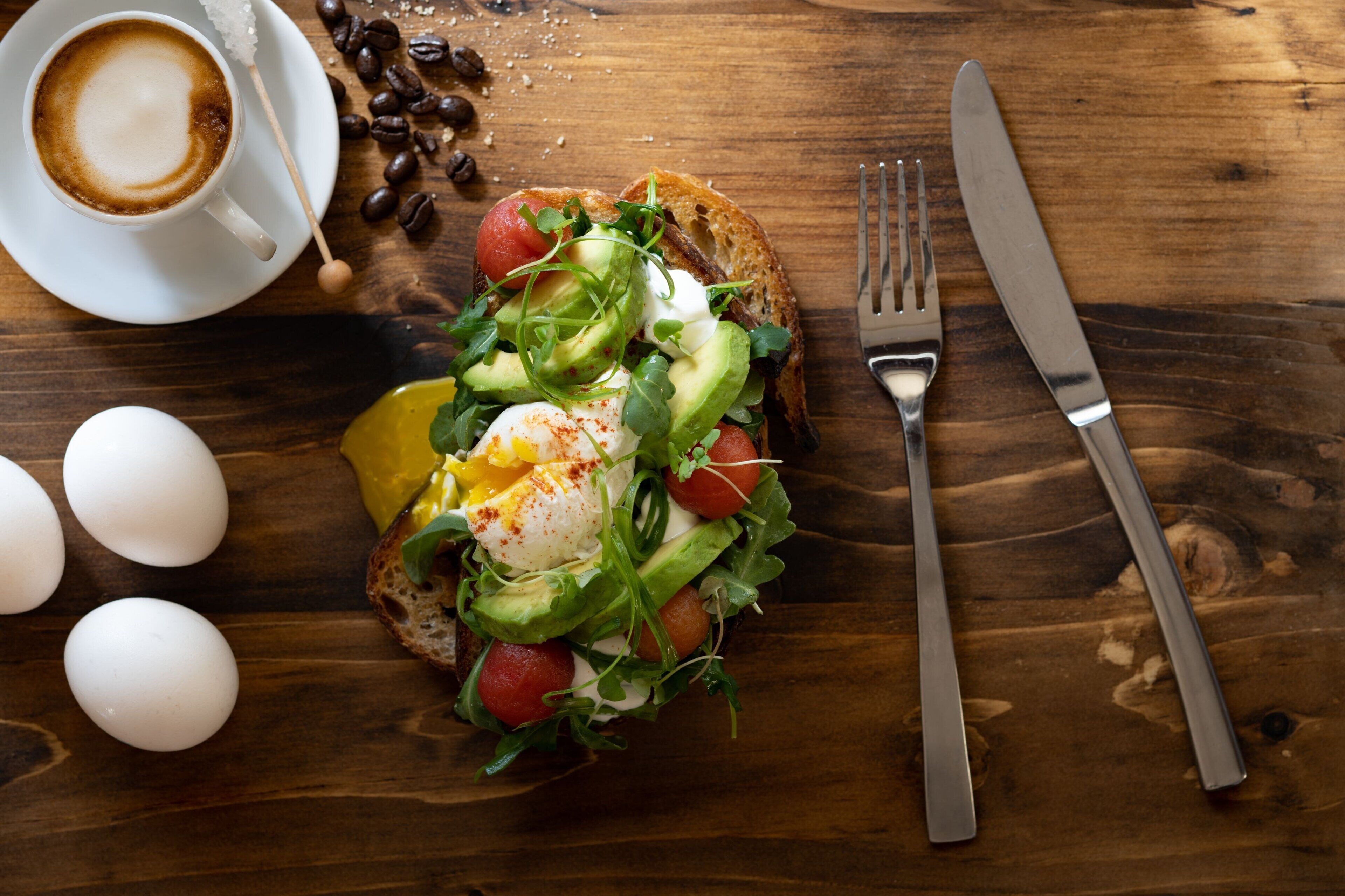 Een ontbijtbord met eieren, avocado, tomaten, bladgroenten op toast, een kop koffie, en een vork en mes op een houten tafel.