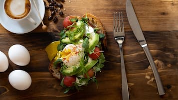 Een ontbijtbord met eieren, avocado, tomaten, bladgroenten op toast, een kop koffie, en een vork en mes op een houten tafel.