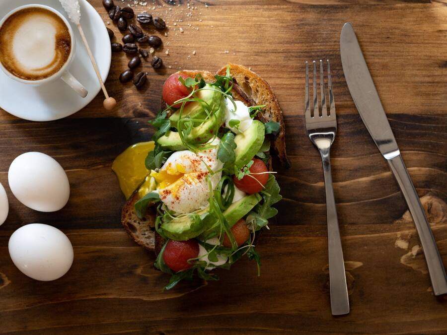 Een ontbijtbord met eieren, avocado, tomaten, bladgroenten op toast, een kop koffie, en een vork en mes op een houten tafel.