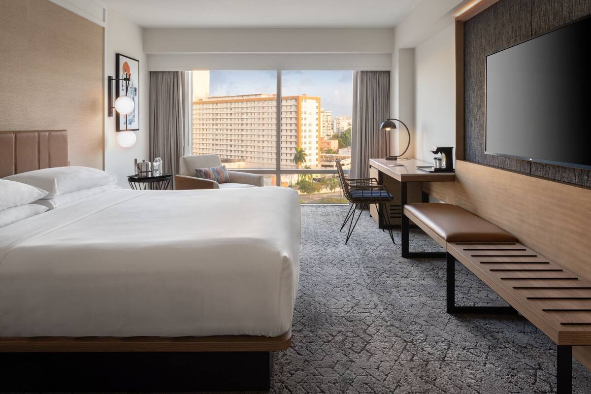 Une chambre d’hôtel avec un grand lit, un bureau avec une chaise, un banc, une télévision et une vue sur le paysage urbain.