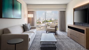 A modern hotel room with a sofa, armchair, coffee table, TV, and large windows offering a city view.