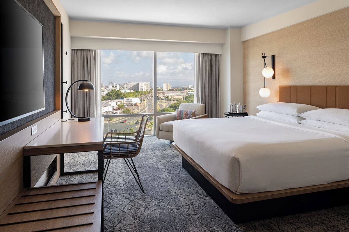 Une chambre d’hôtel avec un grand lit, un bureau avec une chaise, une télévision, une vue sur la ville et une table de chevet avec une lampe.