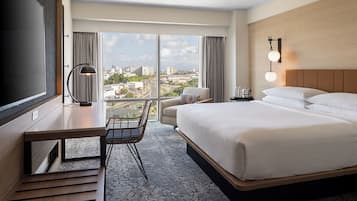 Una camera d'albergo con un letto grande, una scrivania con sedia, una televisione, vista sulla città e un comodino con lampada.