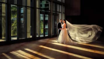 A couple in wedding attire inside a building with large windows and a wooden floor.