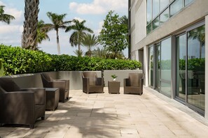 A modern outdoor seating area with wicker furniture, surrounded by lush greenery and palm trees, adjacent to a building with large windows.