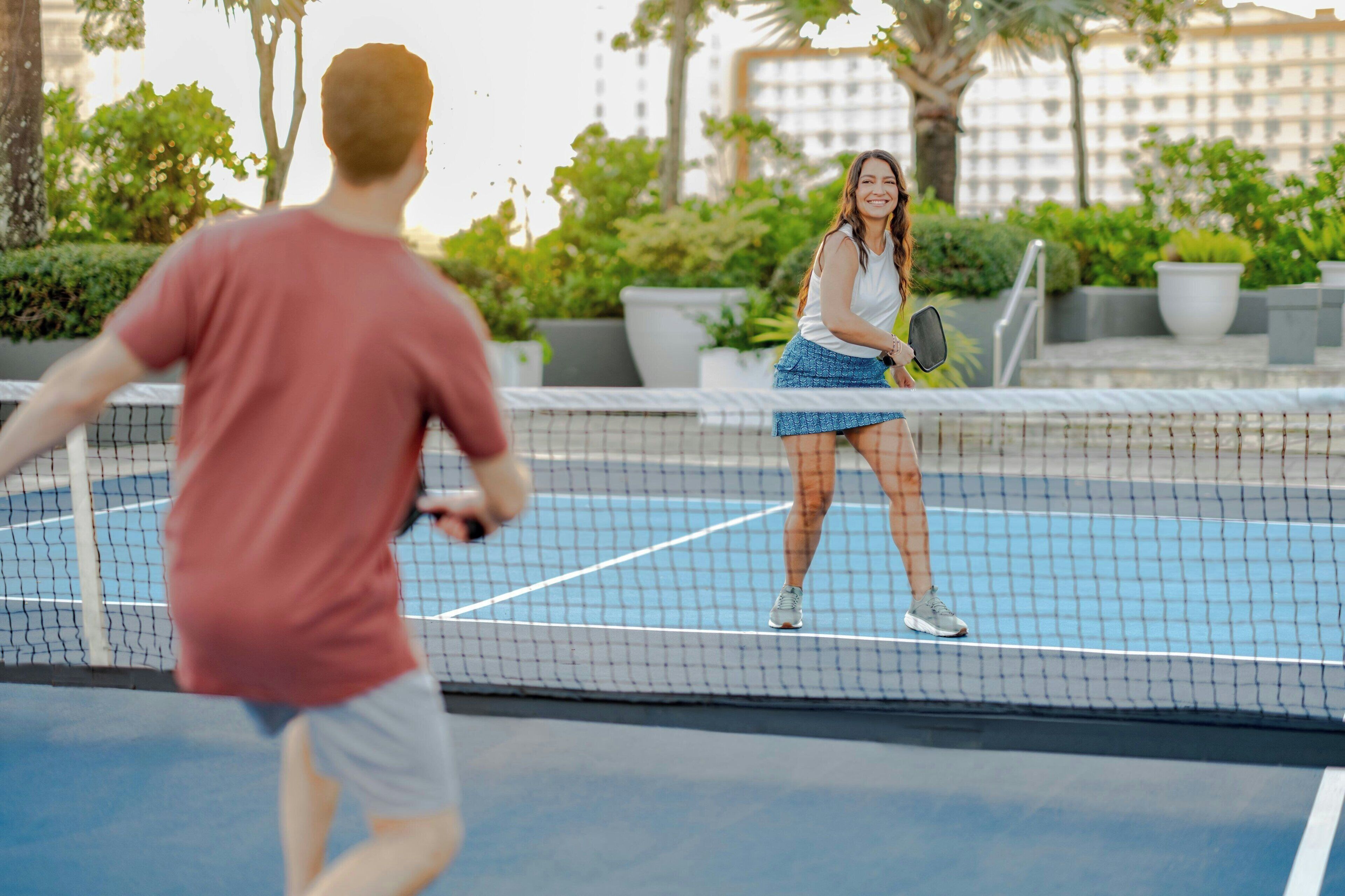 Un court de tennis en plein air, doté d’un filet, entouré de verdure et de bâtiments.
