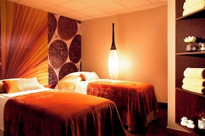 A hotel room with two beds, a stone-patterned wall, a hanging light fixture, and a shelf with towels and a vase.
