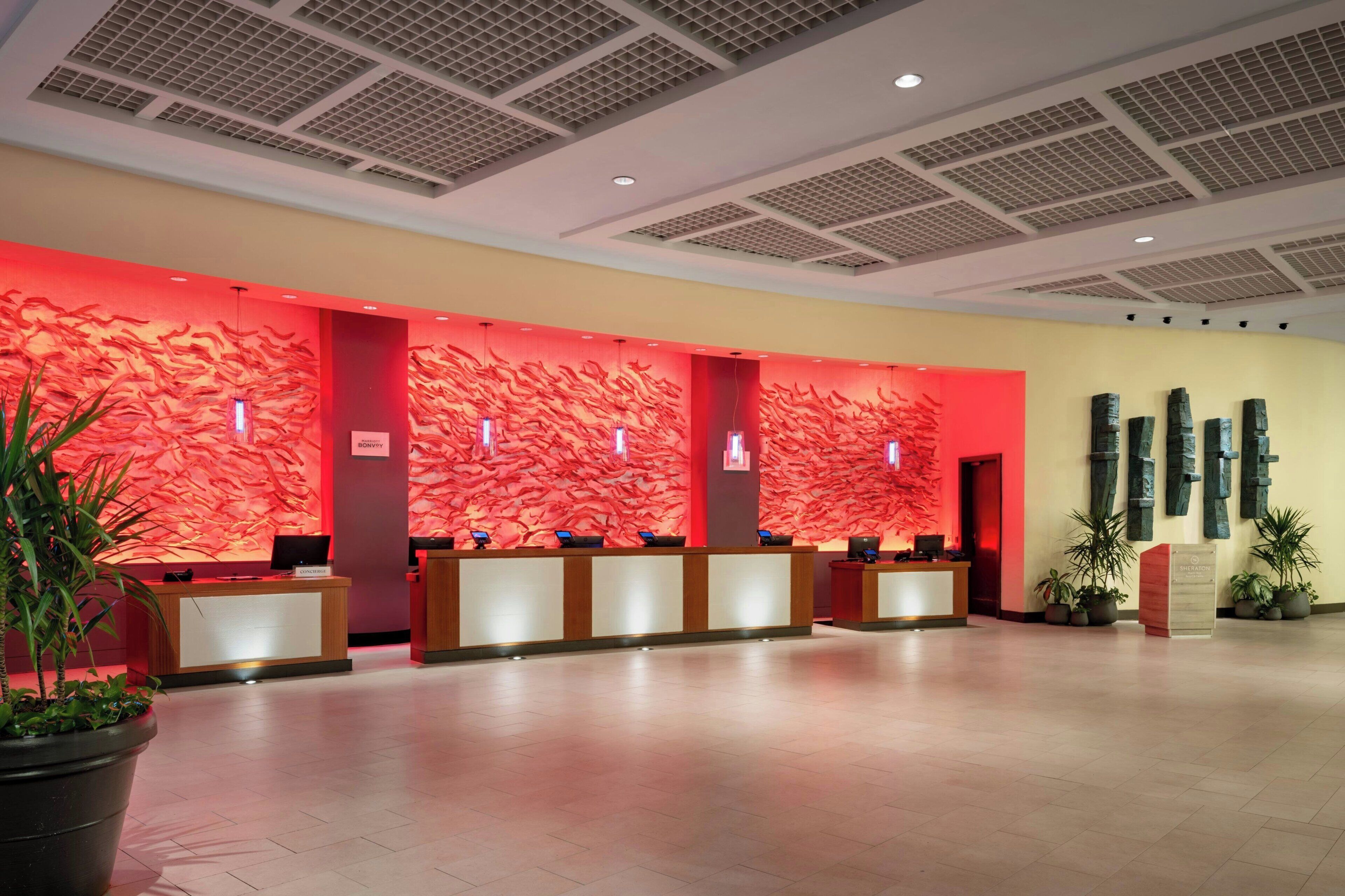 Zona de recepción de un hotel con una pared roja texturizada, varios mostradores de recepción y plantas en macetas.