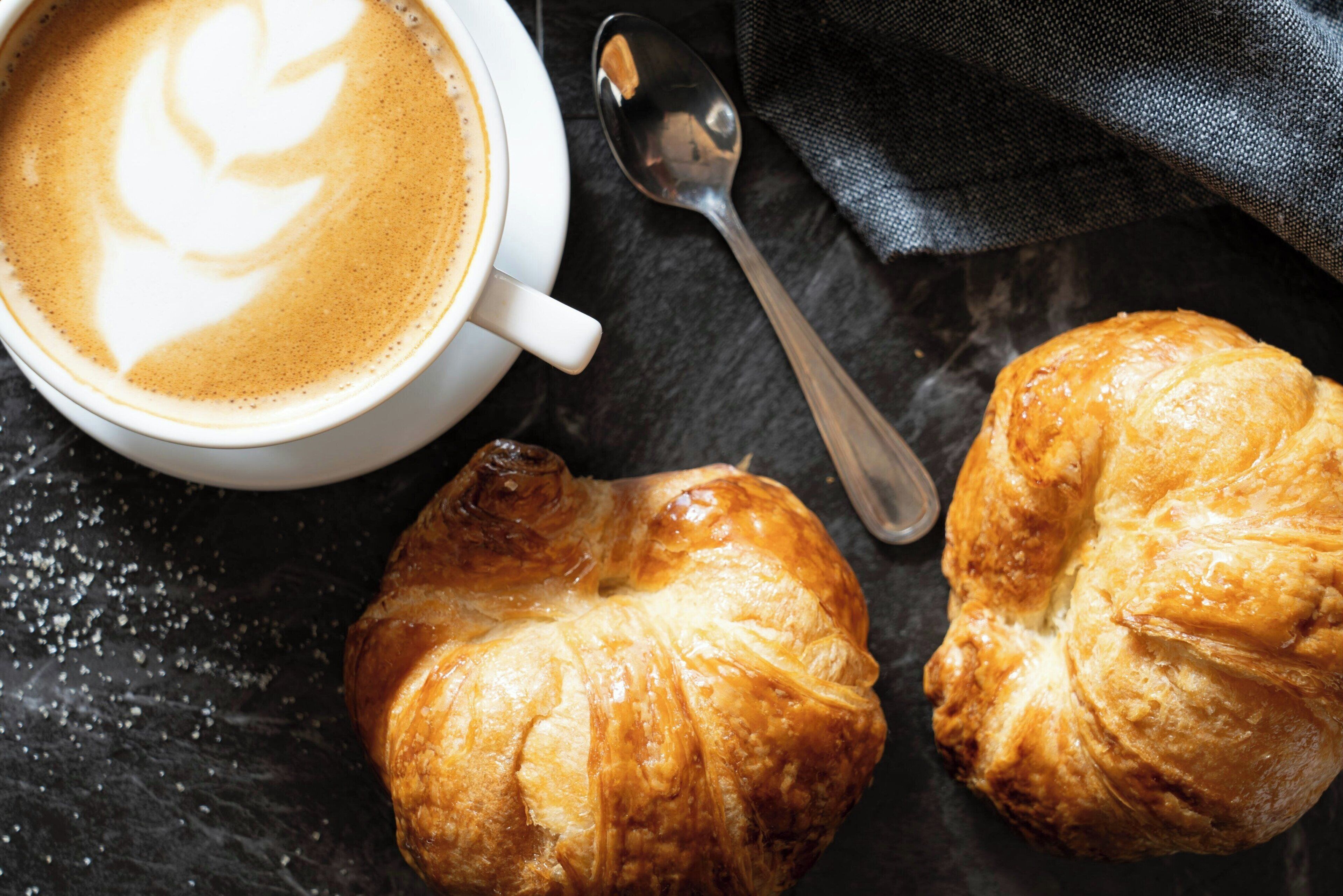 A cup of coffee with latte art and two croissants on a dark surface.