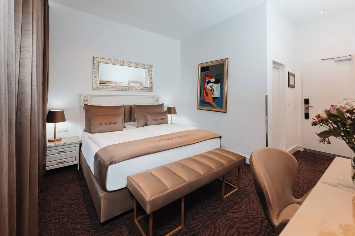 Une chambre d’hôtel avec un grand lit, une banquette, un bureau, une chaise, une table de chevet, une lampe, un miroir et un tableau au mur.