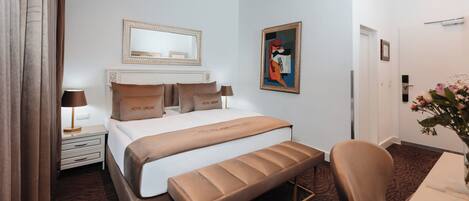 Habitación de hotel con una cama grande, un banco, un escritorio, una silla, una mesita de noche, una lámpara, un espejo y un cuadro en la pared.