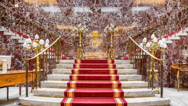 Un magnifique escalier avec des murs de marbre, des marches recouvertes de moquette rouge et d’élégants lustres.