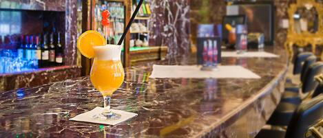 Un bar con encimera de mármol, una cerveza con rodaja de naranja y una pajita, y una estantería con botellas al fondo.