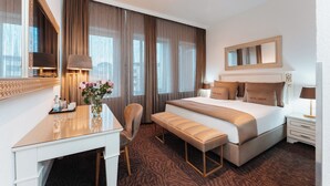 Une chambre d’hôtel avec un grand lit, un bureau, une chaise, un banc, un miroir et une fenêtre avec des rideaux.
