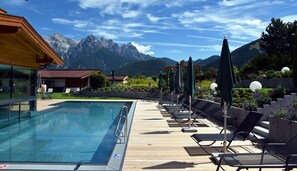 A pool area with lounge chairs and umbrellas, a wooden deck, and a mountain view in the background. - Naturhotel Kitzspitz (Sankt Jakob in Haus)