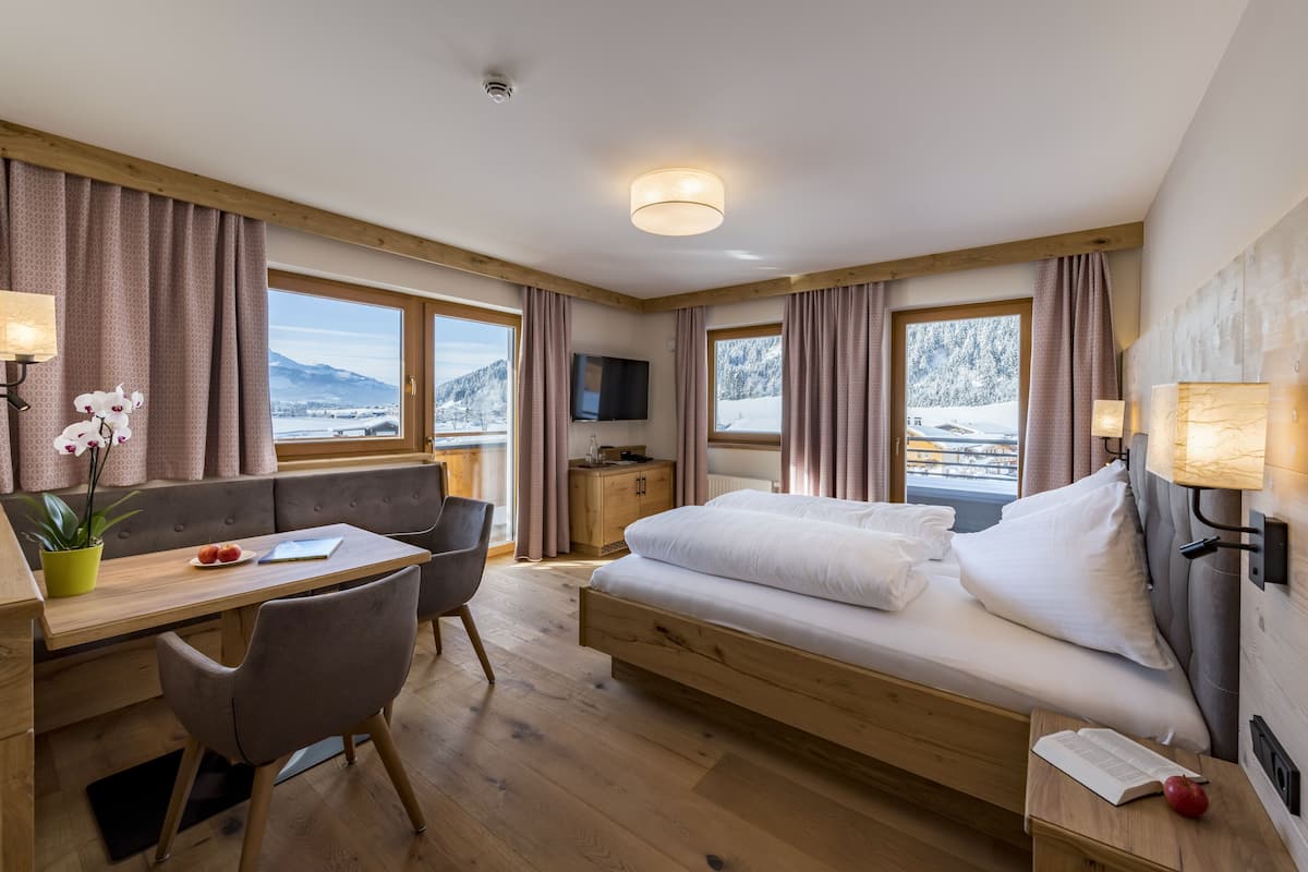 A modern hotel room with a wooden bed, a desk with a chair, a small table with a plant, and a view of snow-covered mountains through large windows.