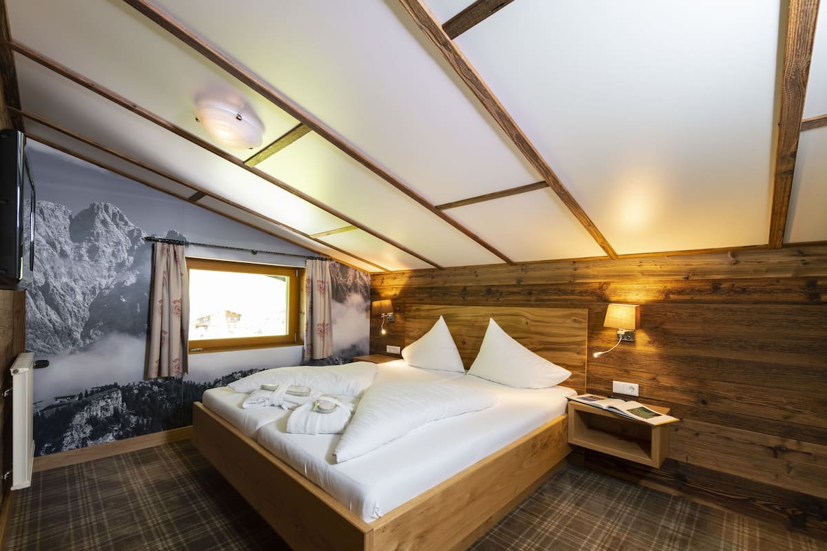 A cozy bedroom with a wooden headboard, a large bed with white linens, a window with curtains, and a wall-mounted lamp.