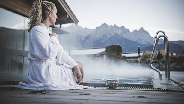 A person in a white bathrobe sitting by a hot tub with a mountainous backdrop. - Naturhotel Kitzspitz (Sankt Jakob in Haus)