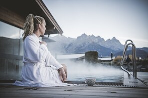 A person in a white bathrobe sitting by a hot tub with a mountainous backdrop. - Naturhotel Kitzspitz (Sankt Jakob in Haus)