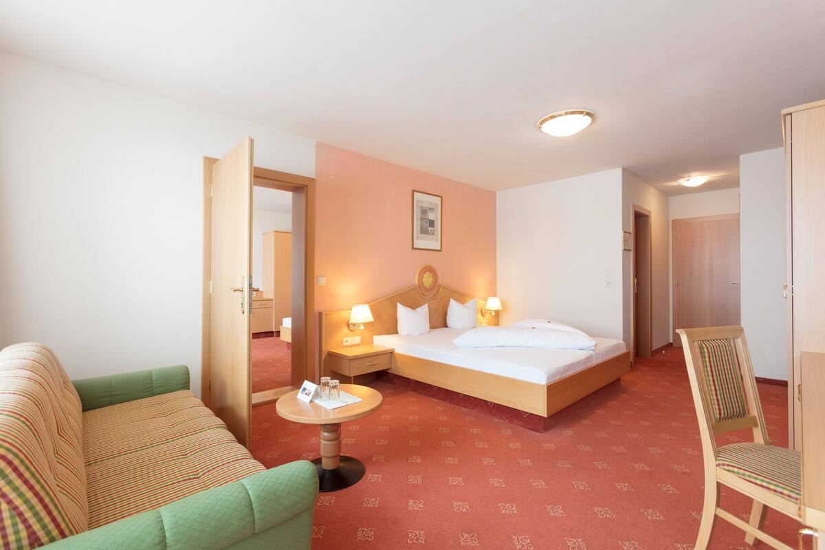A hotel room with a bed, a green sofa, a small round table with a lamp, and a wooden chair.