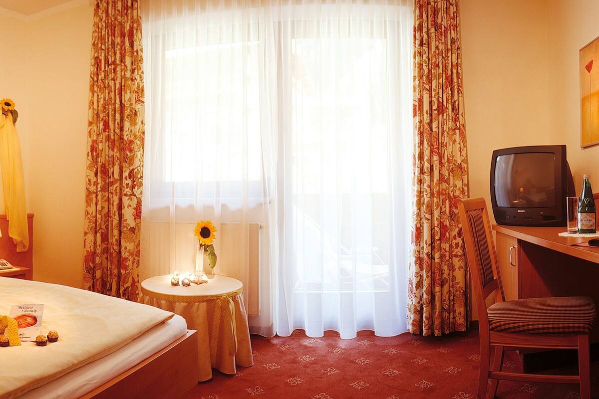 A hotel room with a bed, a desk with a television, a small table with a vase and a sunflower, and curtains.