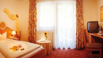 Ein Hotelzimmer mit einem Bett, einem Schreibtisch mit Fernseher, einem kleinen Tisch mit einer Vase und einer Sonnenblume sowie Vorhängen.