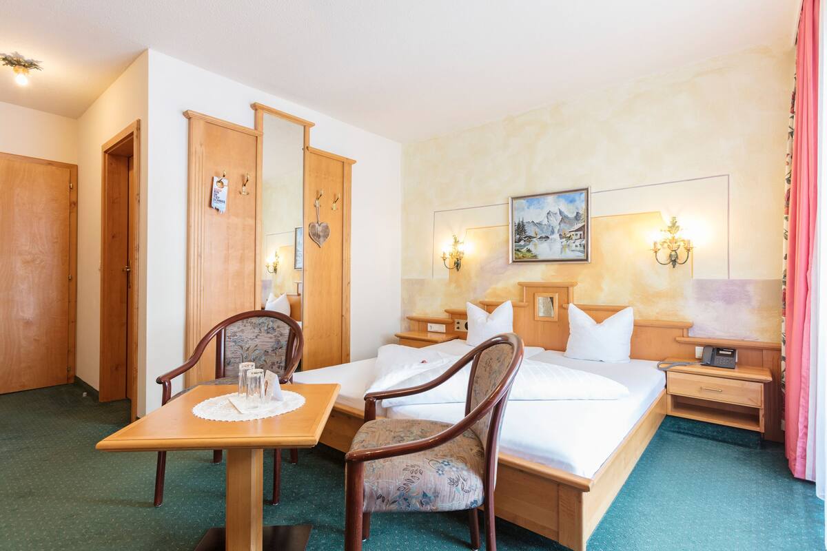 A hotel room with a bed, a wooden nightstand, a table with a chair, and a mirror.