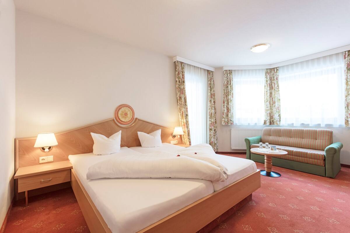 A hotel room with a large bed, a green sofa, a small table, and a nightstand with a lamp.