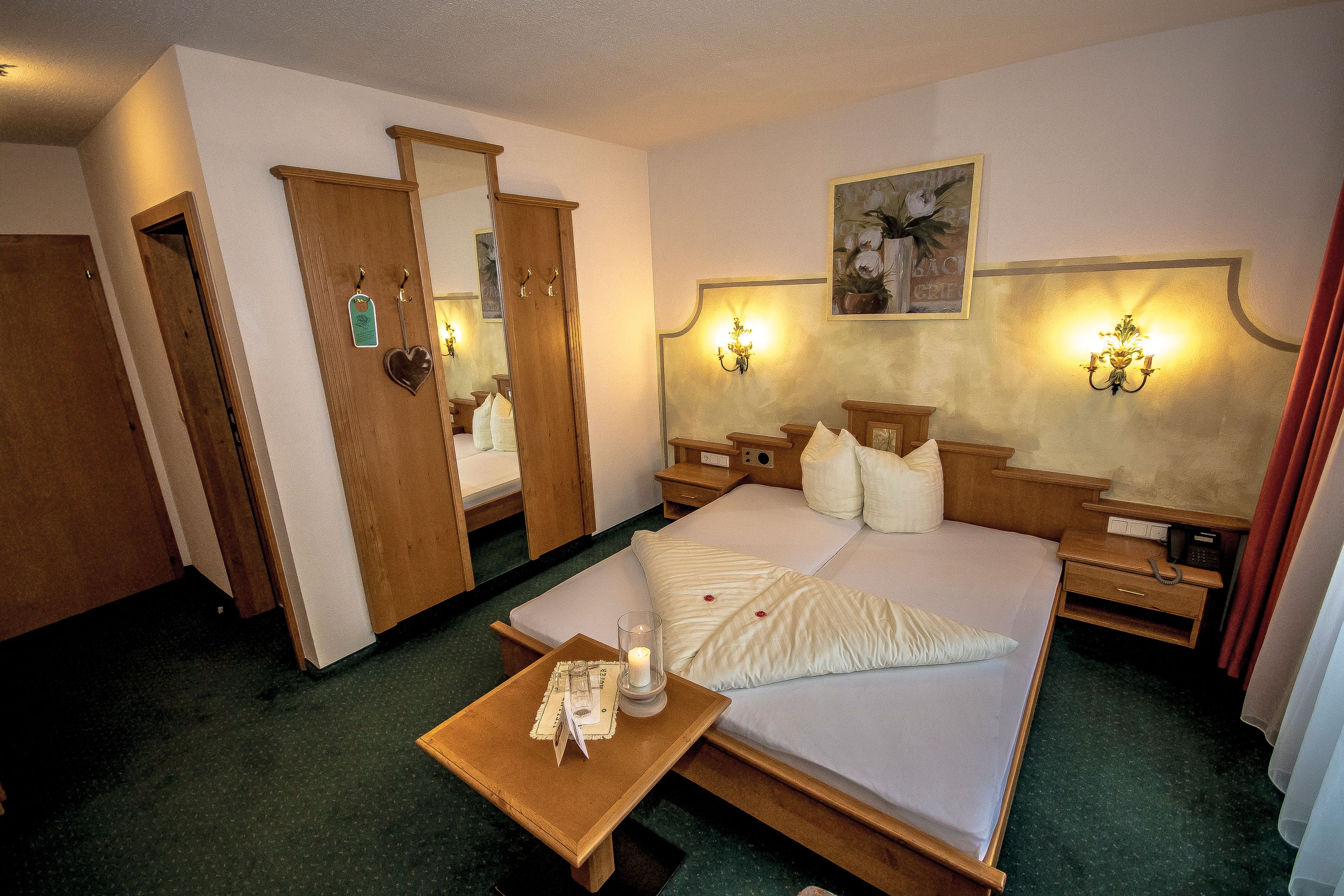 A hotel room with a large bed, wooden furniture, a bedside table with a candle, and wall-mounted lamps.
