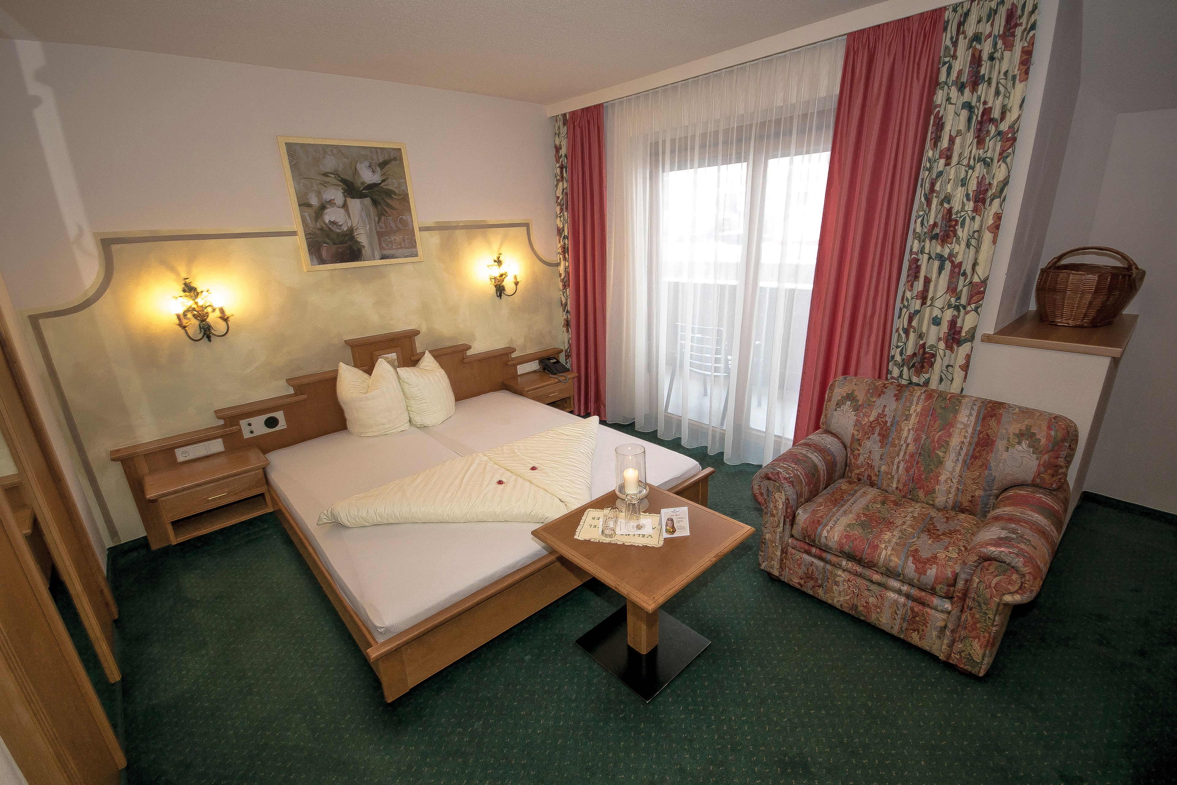 A hotel room with a bed, bedside tables, a nightstand, a chair, a small table with a candle, and a window with curtains.