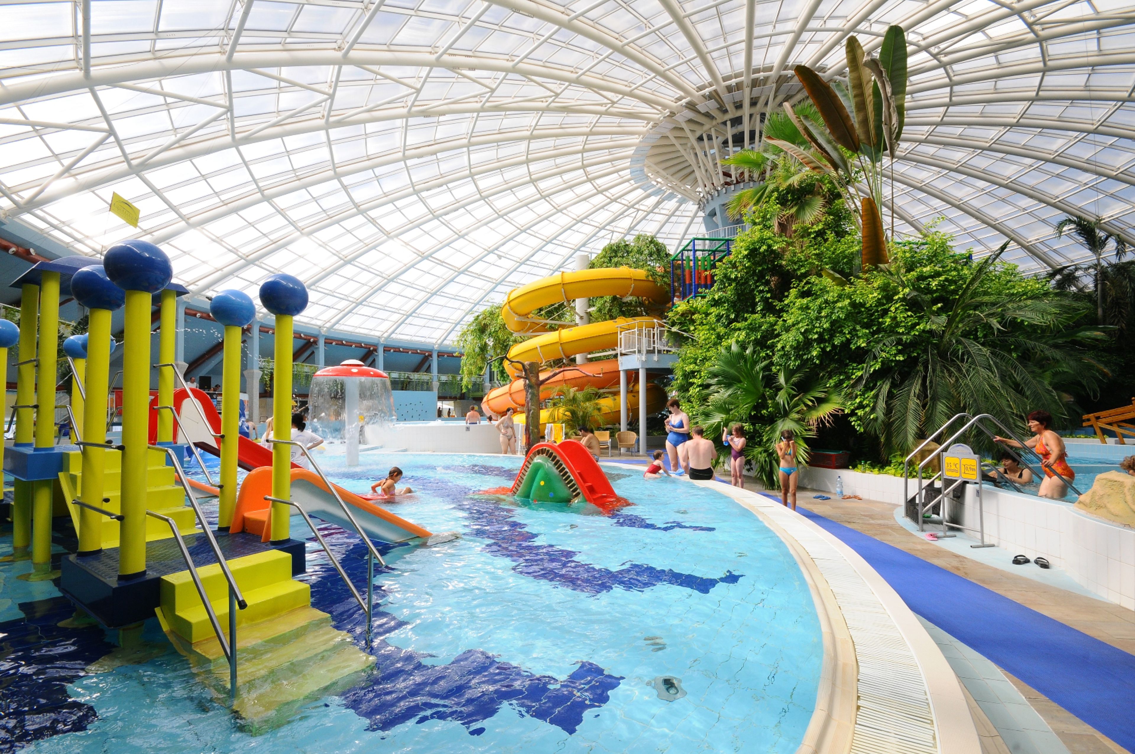 Ein Innenpool mit Rutschen und Wasserspielen, umgeben von üppiger Bepflanzung und einer Glasdecke.