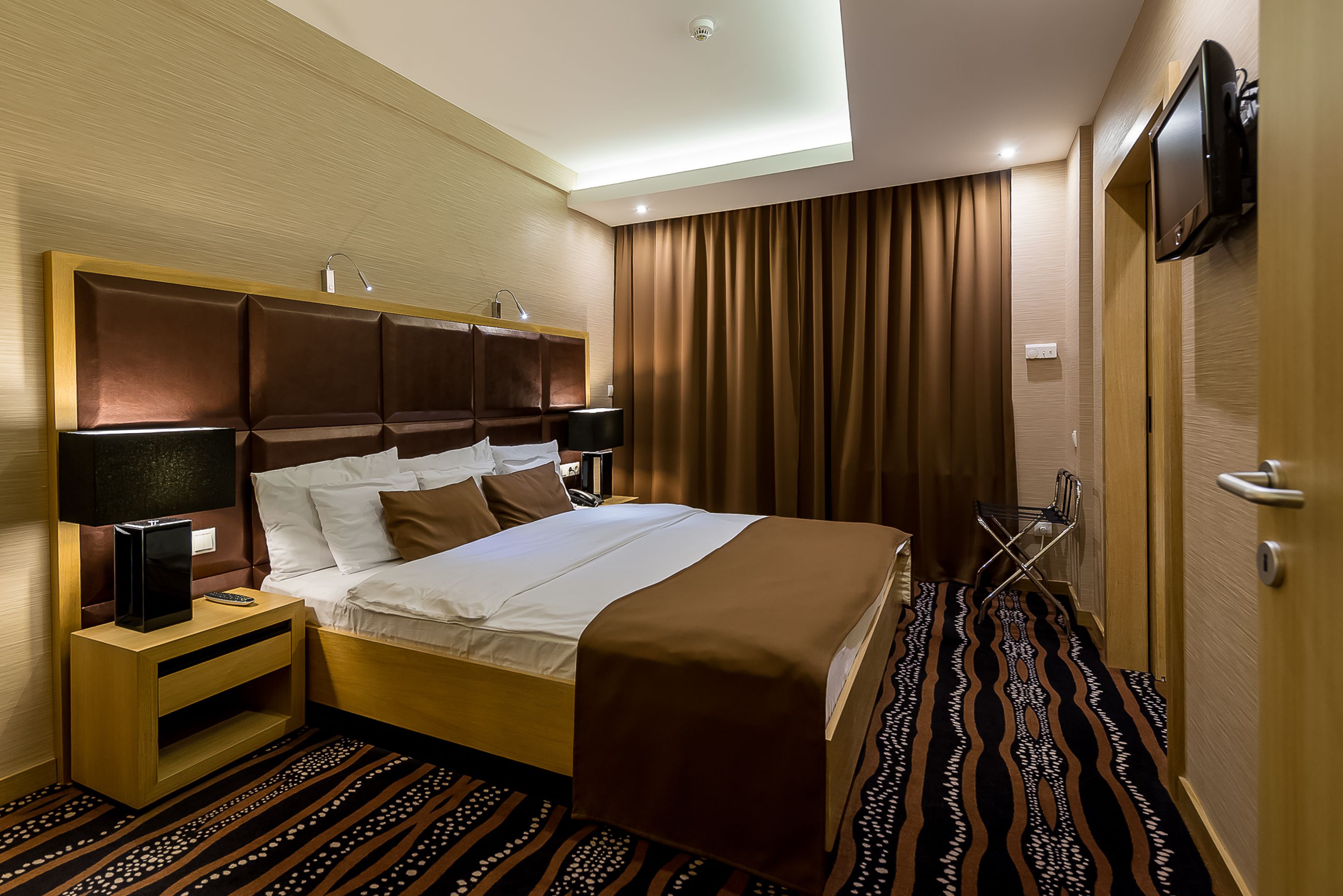 Una habitación de hotel con cama, mesitas de noche, televisor y alfombra estampada.