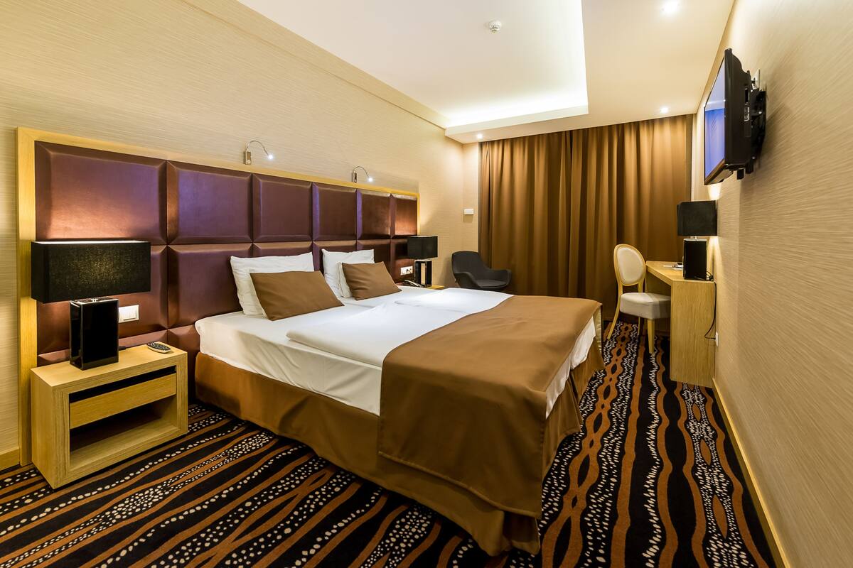 Una habitación de hotel con una cama grande, una mesita de noche con lámpara, un televisor montado en la pared y una alfombra estampada.