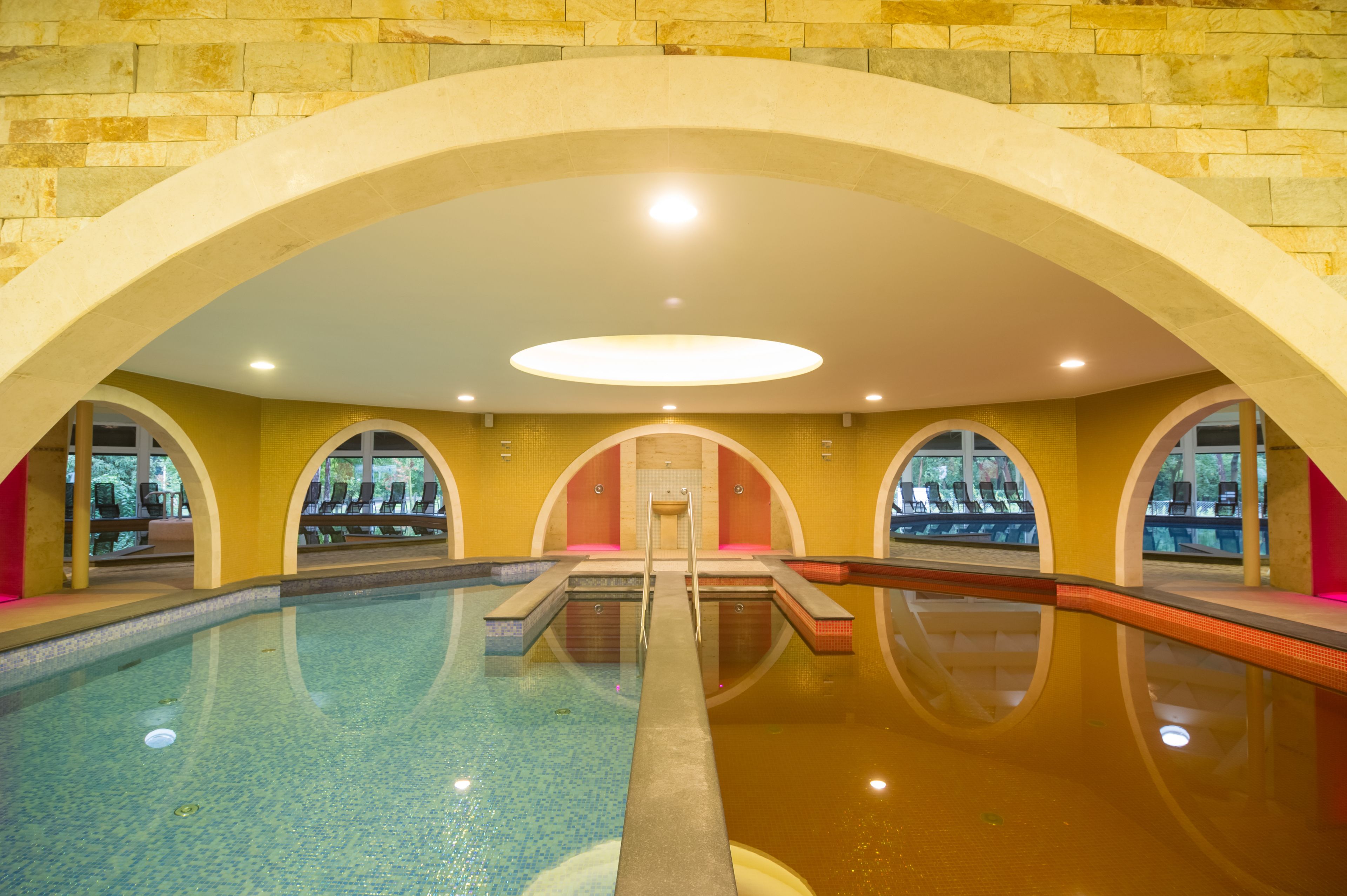 An indoor swimming pool with clear water, a central diving platform, and multiple seating areas with chairs and umbrellas.