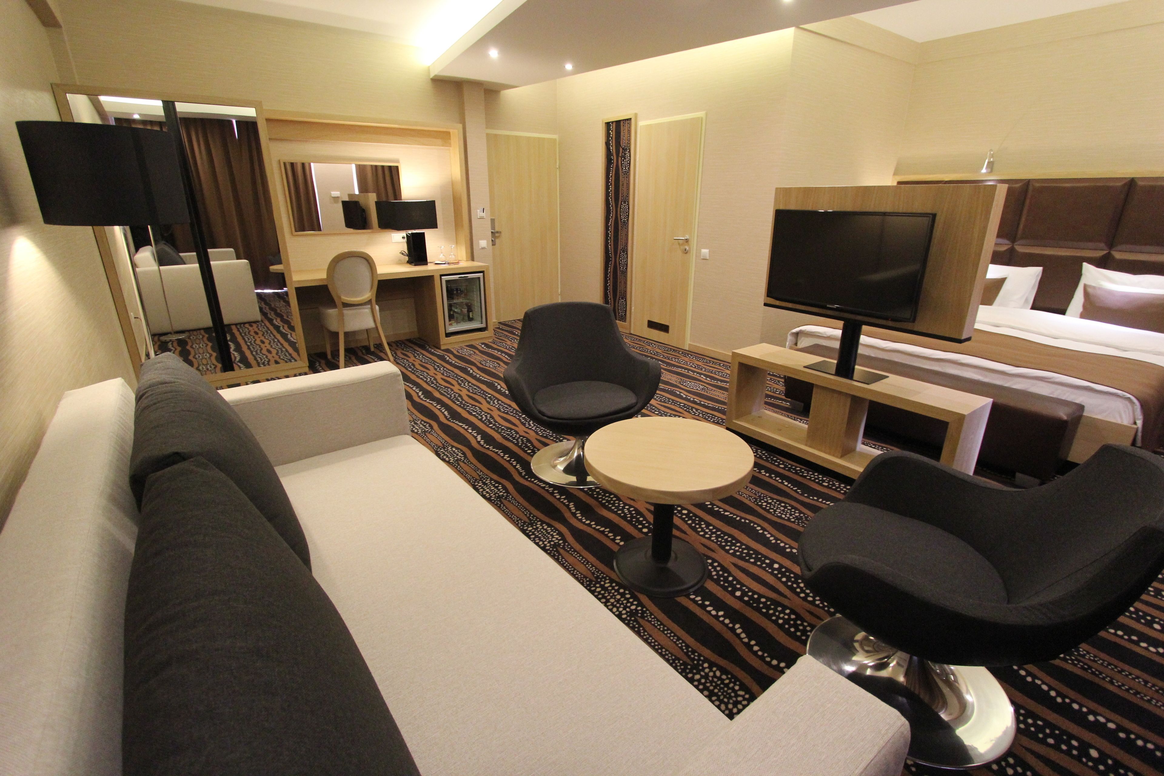 Une chambre d’hôtel comprenant un canapé, deux fauteuils, une petite table ronde, une télévision, un bureau avec une chaise et un miroir.