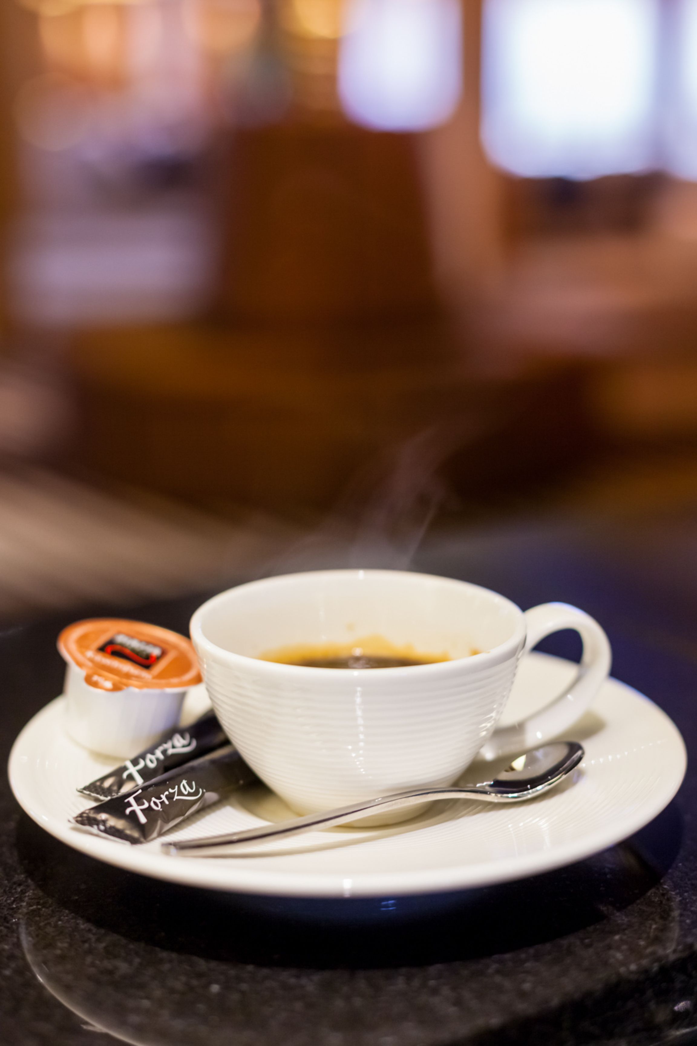 A white cup of coffee on a saucer with a spoon, a small container labeled "Forza," and a small packet on the table.