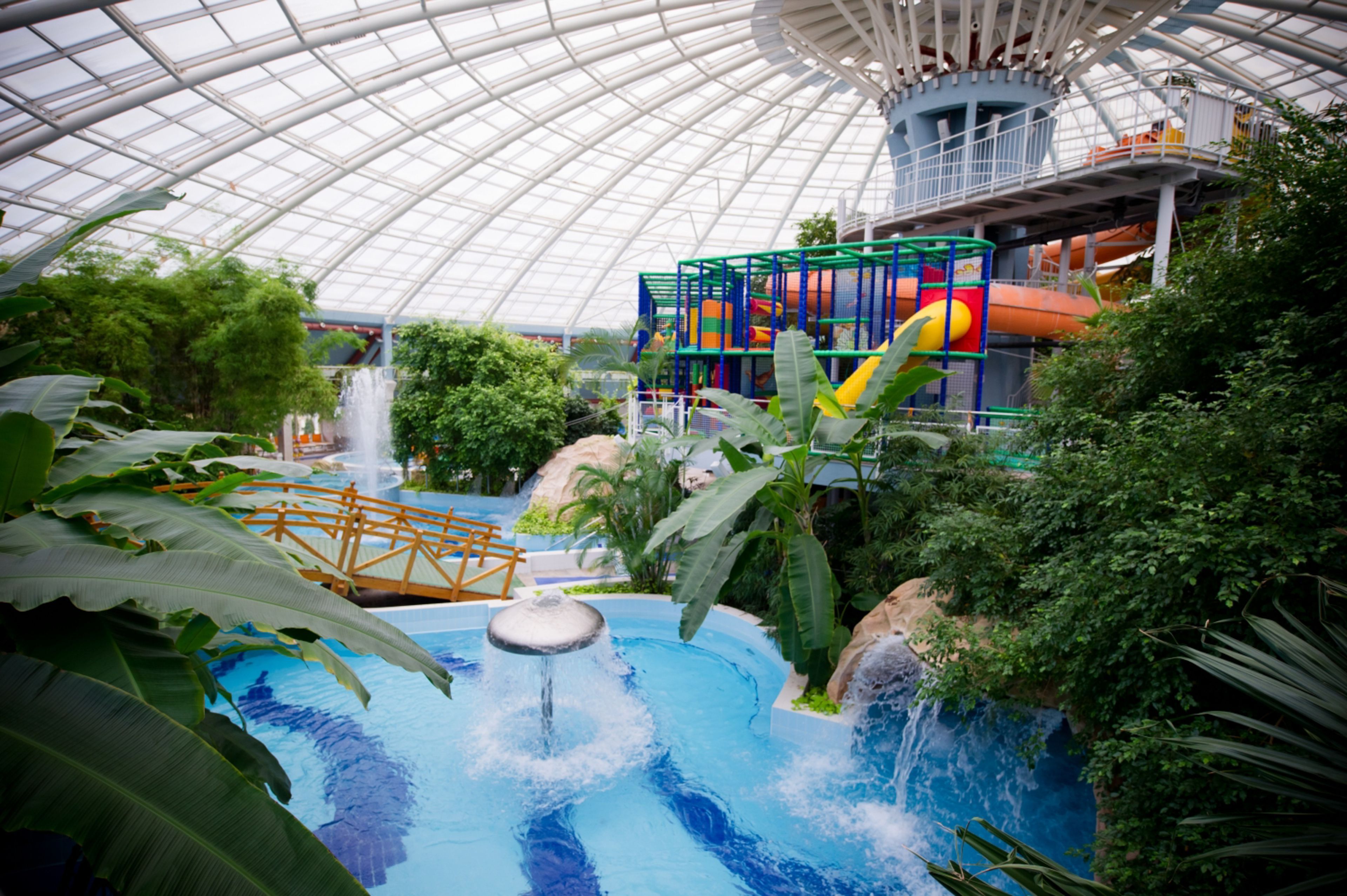 An indoor water park with a large transparent dome ceiling, a central water feature, and a colorful play structure.