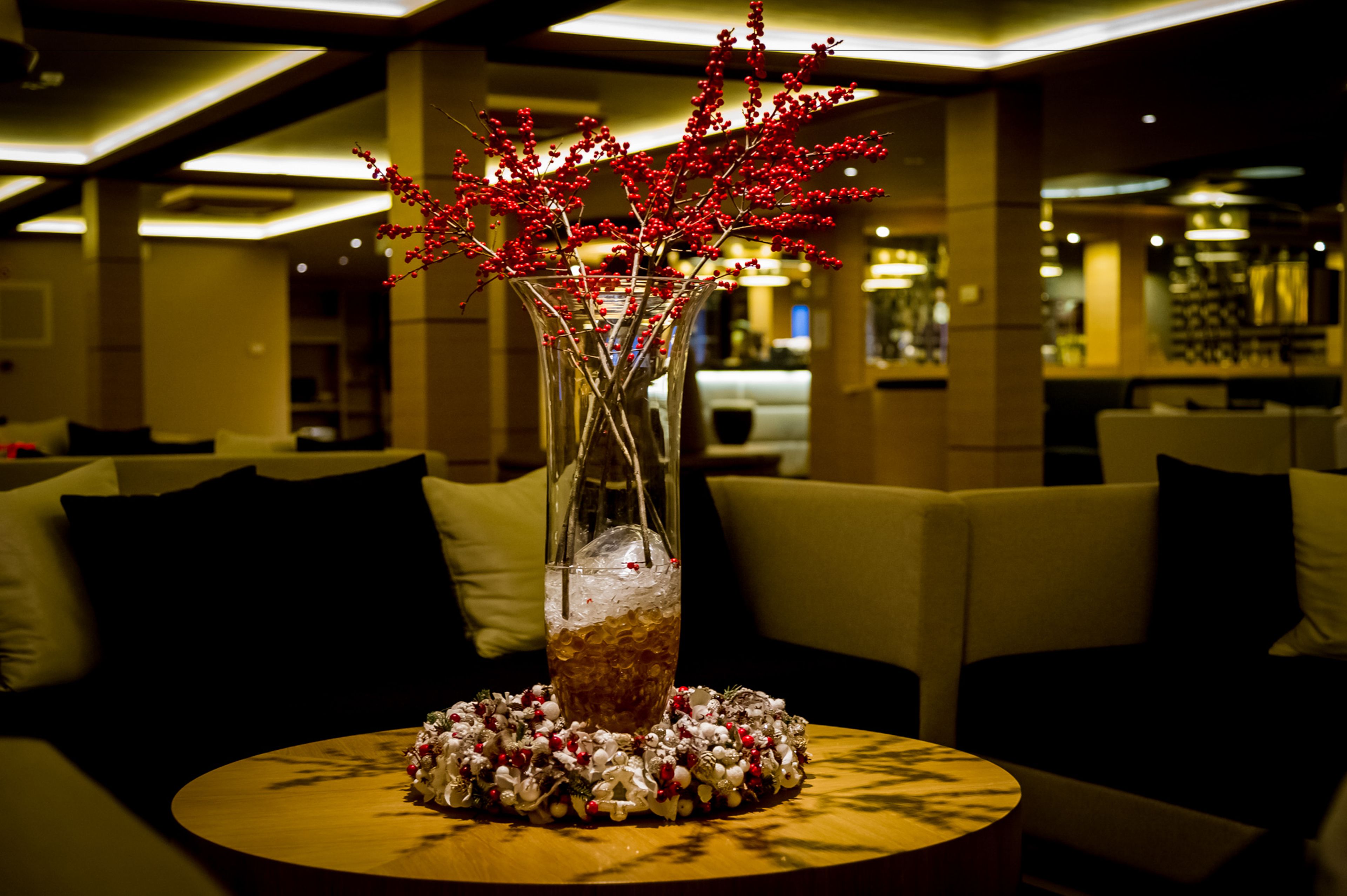 Un tavolo rotondo in legno con un vaso di vetro trasparente contenente bacche rosse e una ghirlanda decorativa, ambientato in una moderna area lounge con divani e un bar sullo sfondo.