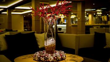 Un tavolo rotondo in legno con un vaso di vetro trasparente contenente bacche rosse e una ghirlanda decorativa, ambientato in una moderna area lounge con divani e un bar sullo sfondo.