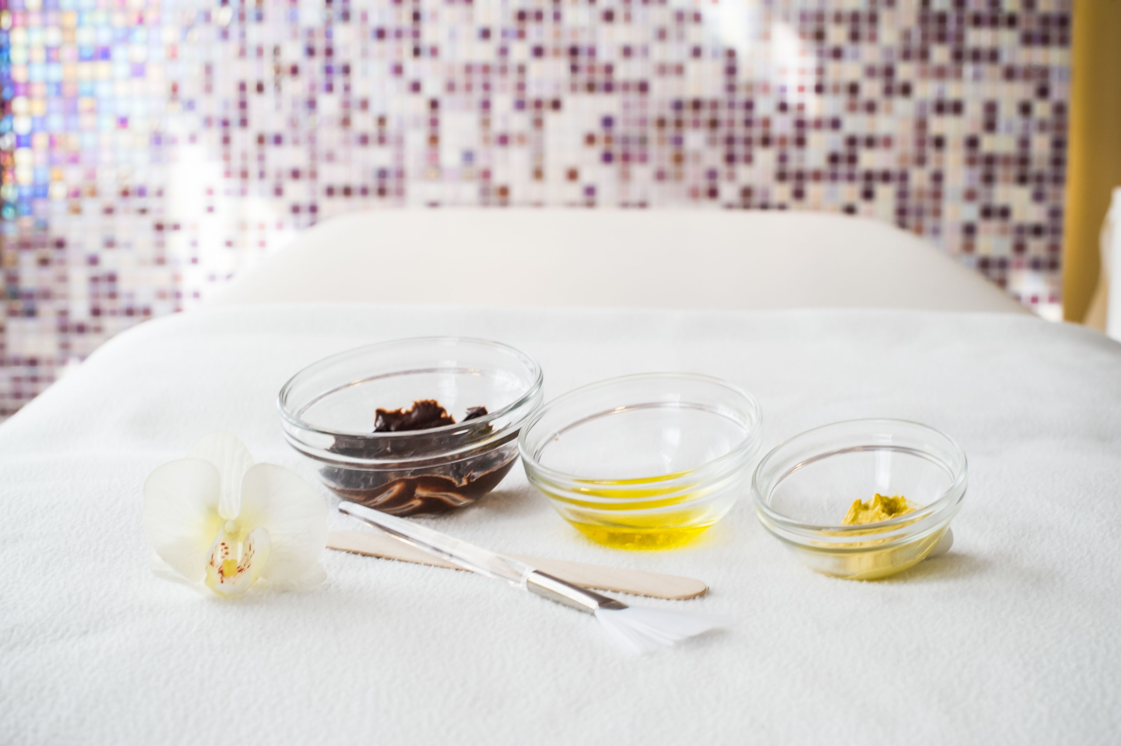 A bed with three glass bowls containing different substances, a white orchid, and a pair of white-handled scissors on a white surface.
