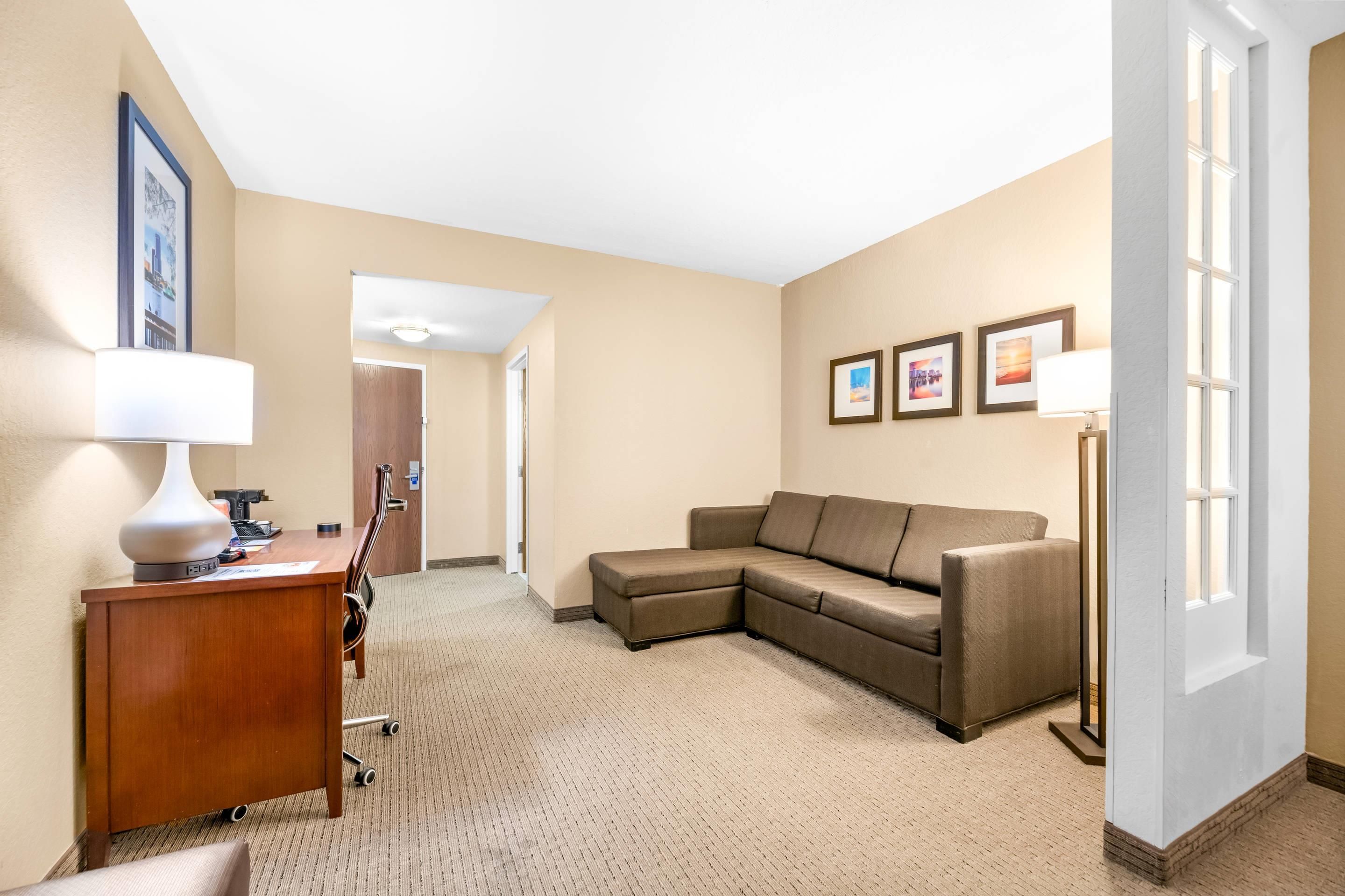 A hotel room with a wooden desk, a brown leather sofa, a standing lamp, and framed pictures on the wall.