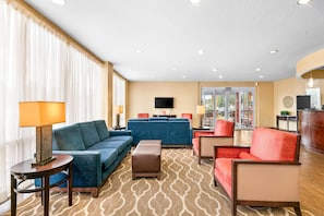 Un hall de hotel con un sofá azul, un sillón rojo, una alfombra estampada y un televisor en la pared.