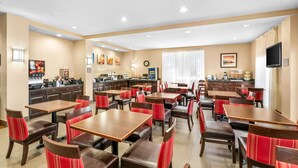 Een restaurantruimte in een hotel met meerdere tafels en stoelen, een keuken op de achtergrond en een televisie aan de muur.