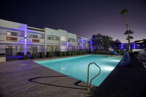 A swimming pool with a metal ladder, surrounded by a landscaped area with trimmed bushes and a palm tree. - Los Viajeros Inn (Wickenburg)