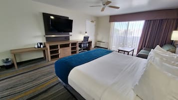 A hotel room with a large bed, a flat-screen TV, a desk with a chair, a small table, and a ceiling fan.