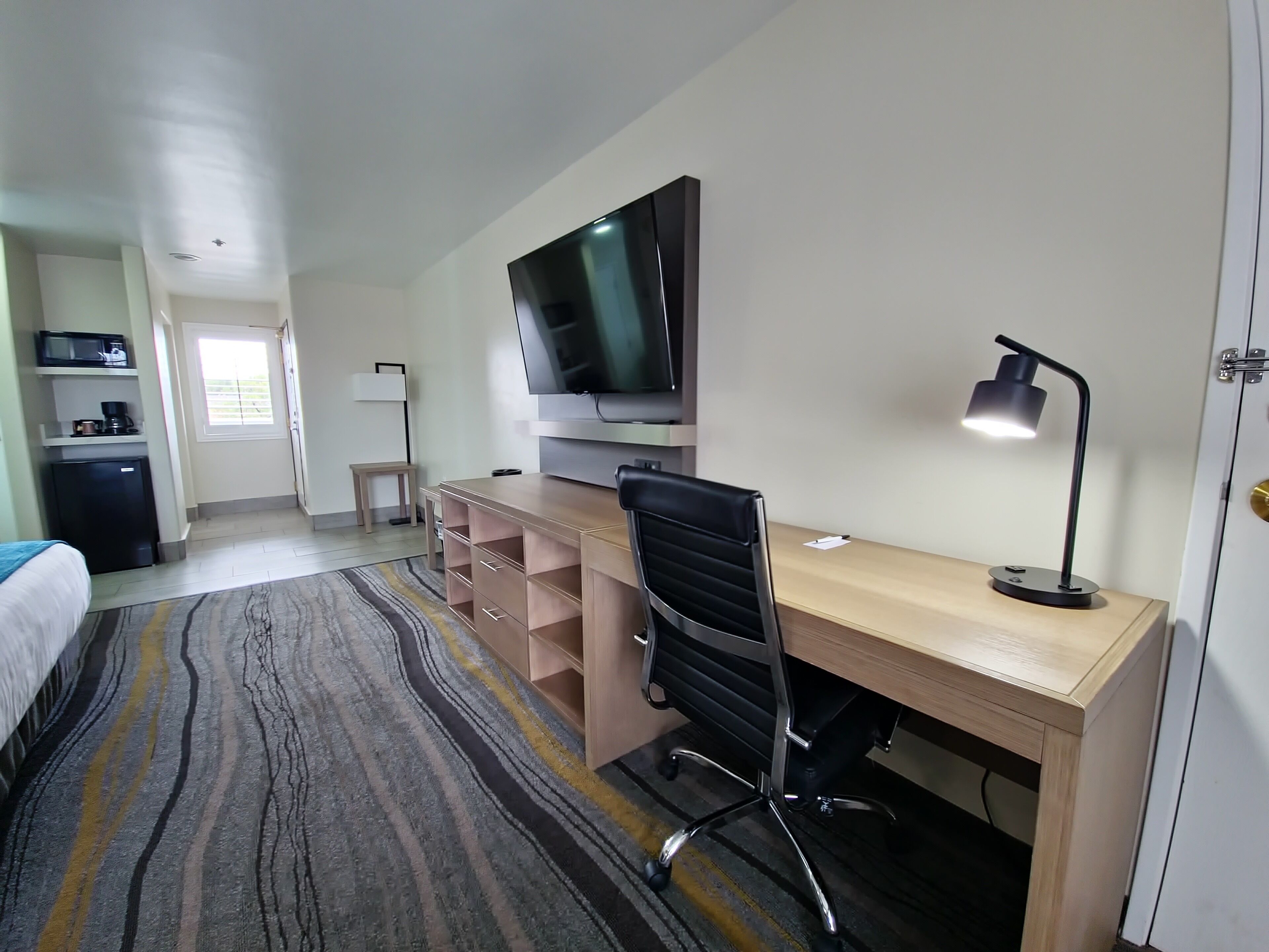 Et hotelværelse med et træskrivebord, en sort kontorstol, et fladskærms-tv ophængt på væggen, en skrivebordslampe, en mikrobølgeovn, et køleskab og en seng med en stribet måtte.