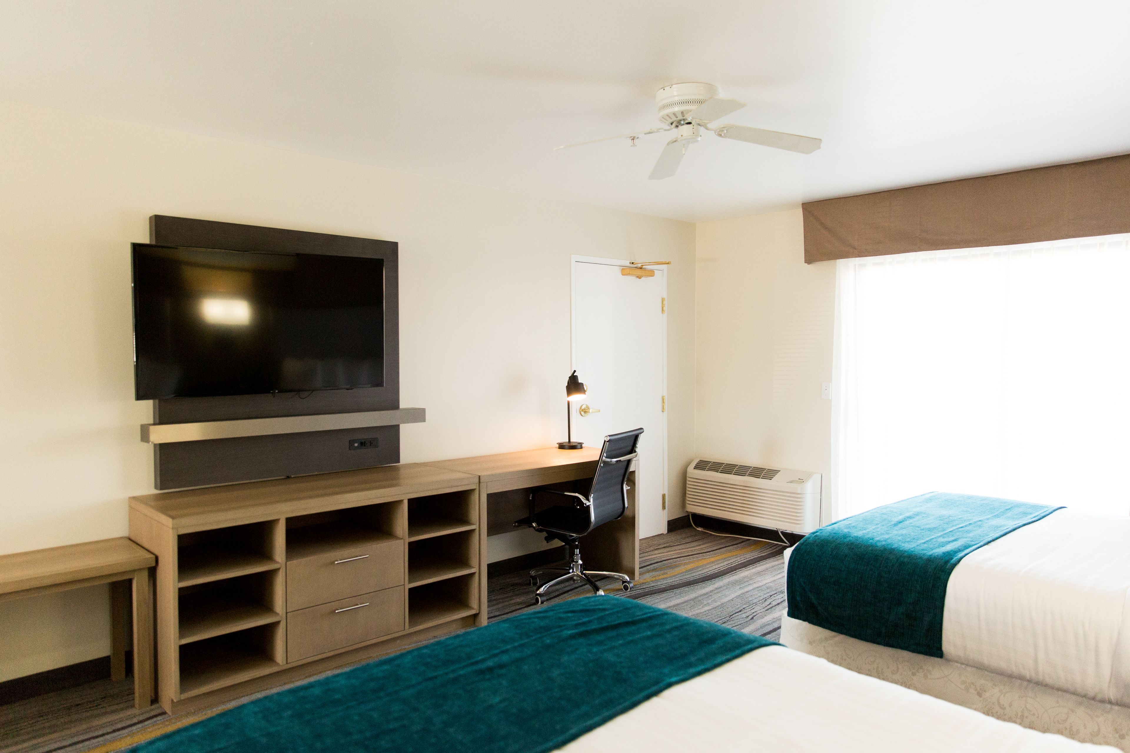 A hotel room with a bed, a desk with a chair, a television, a ceiling fan, and an air conditioning unit.