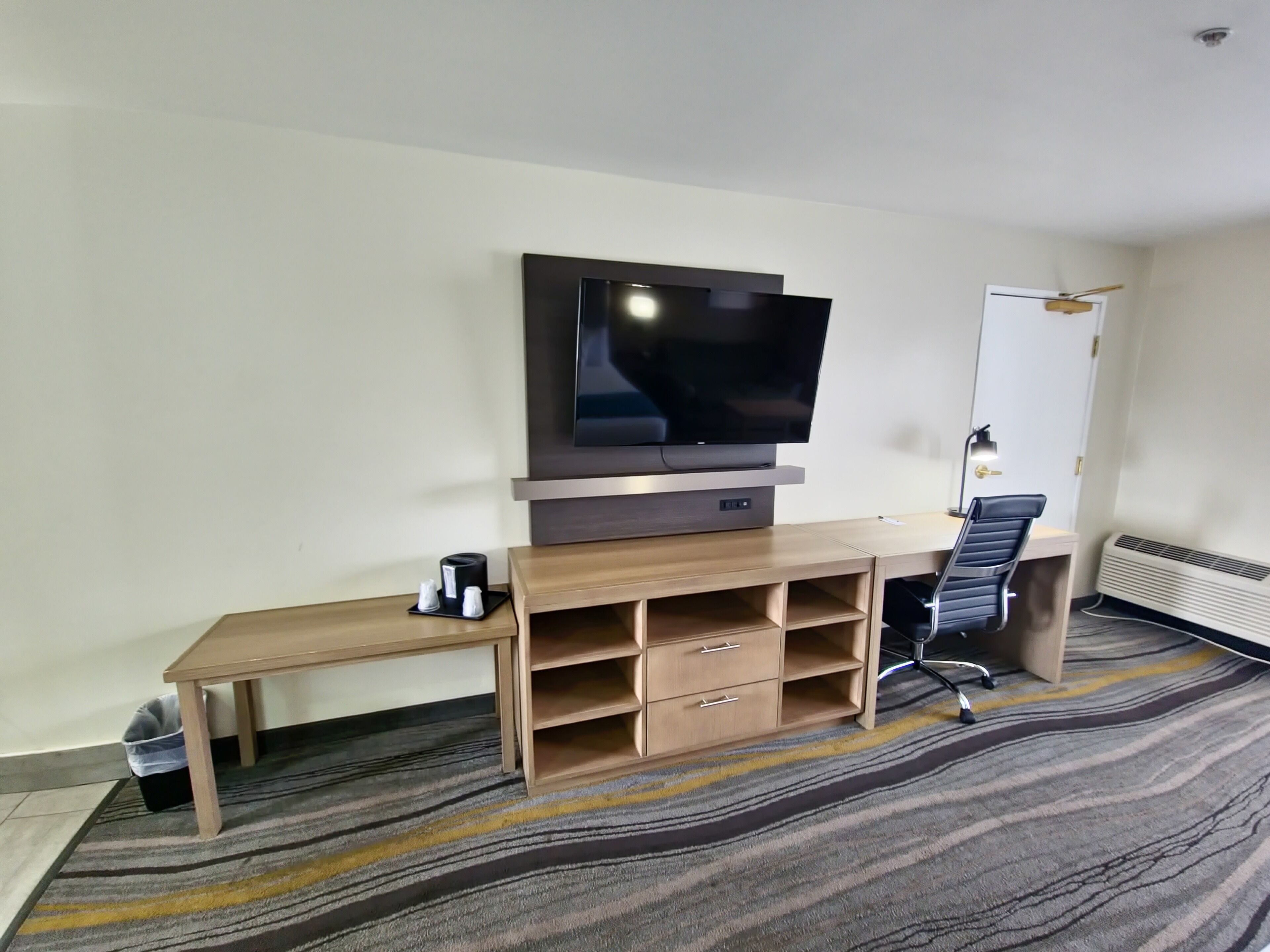 A hotel room with a flat-screen TV mounted on a wooden console, a desk with a chair, and a standing heater.