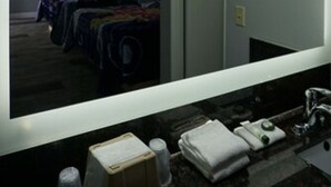 A hotel room with a large mirror, a bed with multiple pillows, a bedside table, and a bathroom with a sink and towel dispenser.