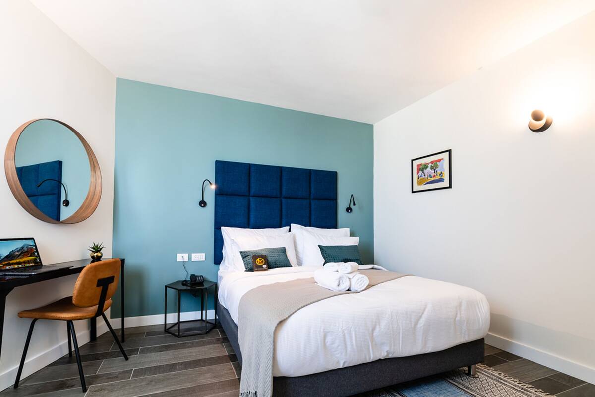 A modern hotel room with a large bed, a desk with a computer, a chair, and a small table with a lamp.